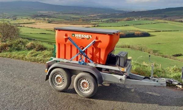 Wessex Polar One Salt And Grit Spreaders | Middlewich Machinery A wessex polar one salt and grit spreader, painted in orange, is parked on the roadside with a vast, green hilly landscape in the background.